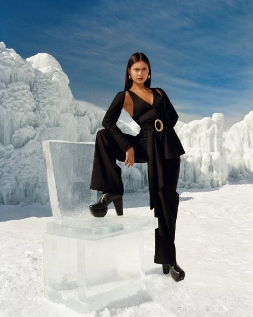 A woman in a black pantsuit poses in a snowy landscape next to a chair made of ice, with snow and a blue sky with white clouds in the backdrop.