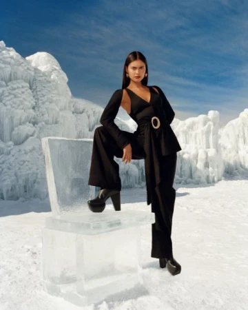 A woman in a black pantsuit poses in a snowy landscape next to a chair made of ice, with snow and a blue sky with white clouds in the backdrop.