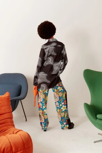 Back view of a woman with an afro hairstyle wearing a patterned shirt and pants with platform shoes, posing in front of a white wall.