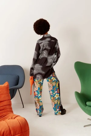 Back view of a woman with an afro hairstyle wearing a patterned shirt and pants with platform shoes, posing in front of a white wall.
