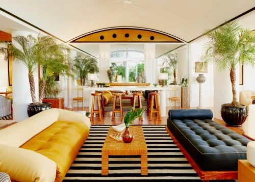 Living room interior with a mustard yellow sofa, a black daybed, striped rug, a coffee table, and potted palm trees.