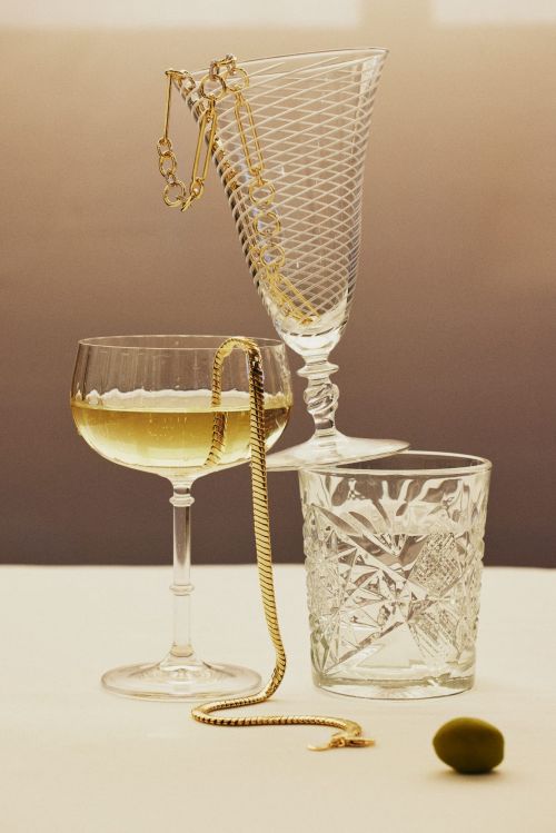 Still life featuring three crystal glasses, a golden chain necklace, and a green olive on a white surface against a gradient background.