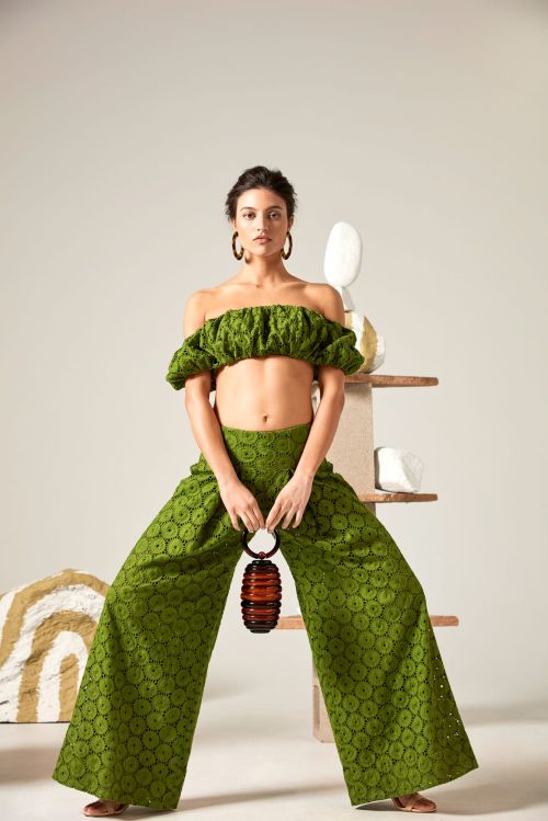 Woman in green two-piece outfit holds a rounded handbag, striking a pose in front of a neutral backdrop with geometric objects.