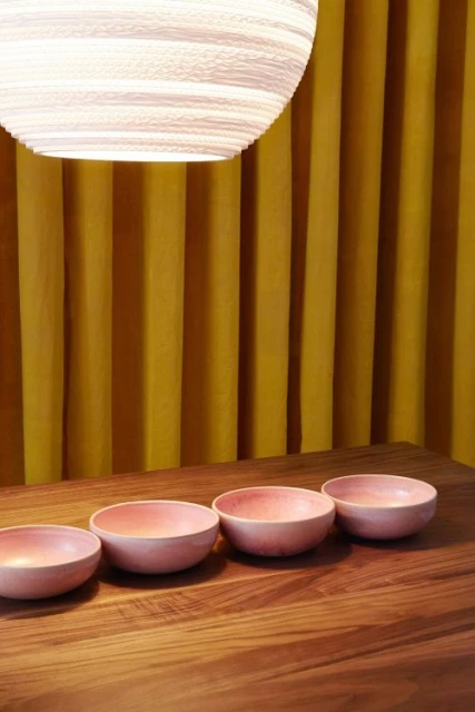 A minimalist still life features four pink ceramic bowls on a wooden table against a vibrant yellow curtain backdrop and modern light fixture.