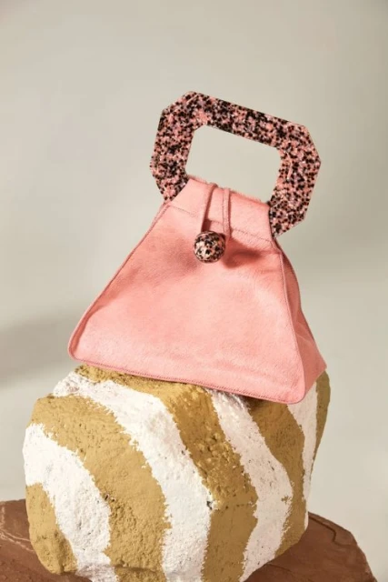 A pink, triangular handbag with a patterned handle sits atop a sculptural, striped block in a fashion studio setting, product-focused.