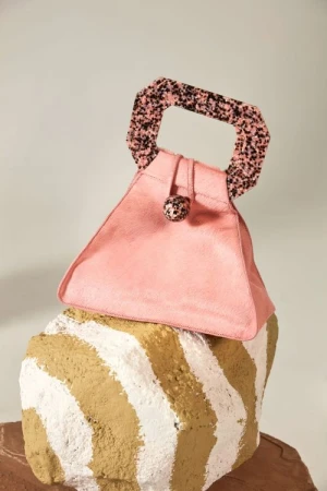 A pink, triangular handbag with a patterned handle sits atop a sculptural, striped block in a fashion studio setting, product-focused.