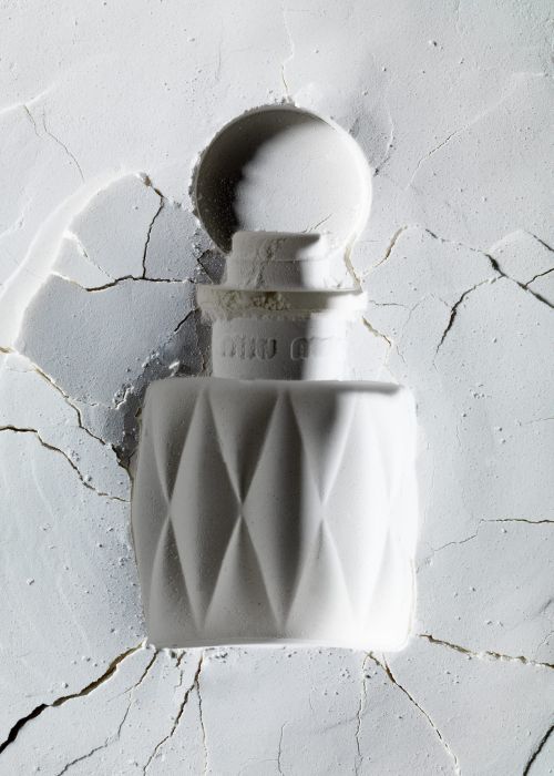 Close-up of a white perfume bottle with a geometric diamond pattern against a textured white powder background in soft, diffused lighting.