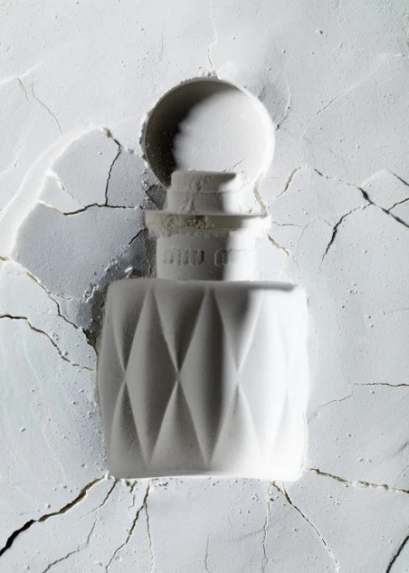 Close-up of a white perfume bottle with a geometric diamond pattern against a textured white powder background in soft, diffused lighting.