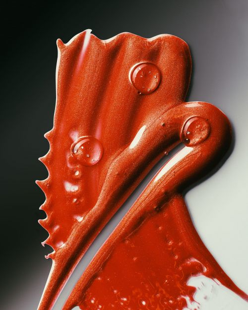 Close-up of a vibrant, shimmering red cosmetic cream swirled on a neutral surface, with water droplets accentuating its glossy texture.