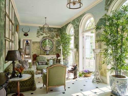 Ornate living room interior with green furnishings, landscape mural wallpaper, large windows, decorative objects, and patterned flooring.