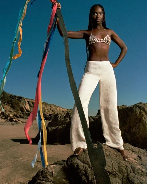 A dark-skinned woman in a bikini top and white pants poses on rocks at the beach, holding flowing colorful ribbons in a fashion editorial.