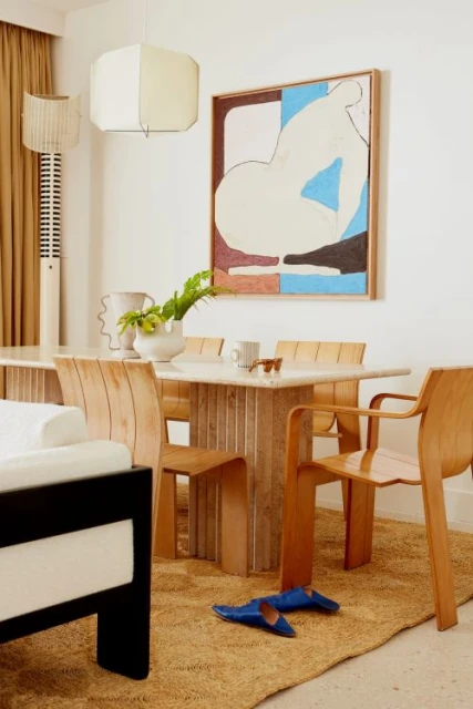 Neutral-toned dining room with travertine table, wooden chairs, art, and a tan rug, illuminated by natural light and modern lighting.