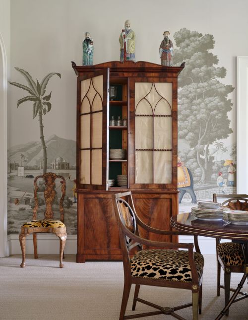 Elegant dining room with a wooden china cabinet displaying dishware, surrounded by decorative chairs and scenic wallpaper.
