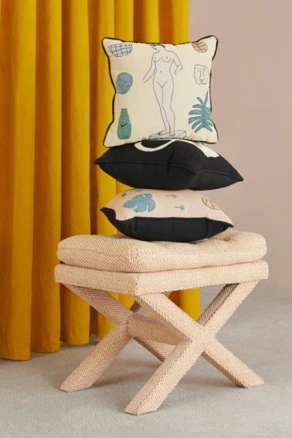 Stack of decorative pillows with various designs sits atop a woven beige stool against a mustard-yellow curtain and light beige wall.