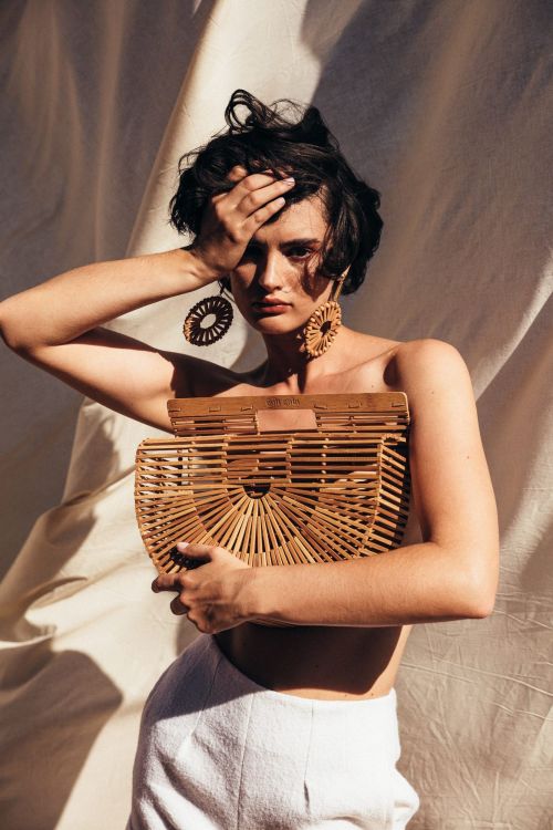Woman with short dark hair, wearing earrings, holding a woven purse against a draped fabric backdrop in soft, diffused lighting.