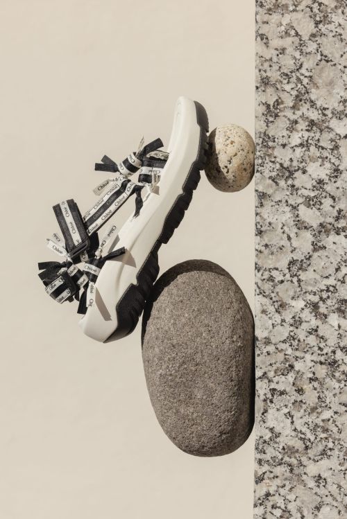 Studio shot of a white sandal with black straps, resting on grey and tan stones, set against a tan and mottled grey background.