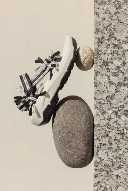 Studio shot of a white sandal with black straps, resting on grey and tan stones, set against a tan and mottled grey background.