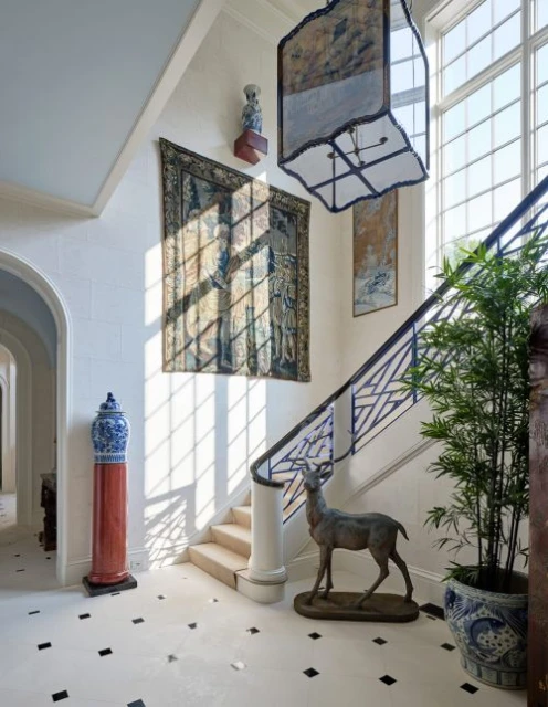 Elegant foyer interior with natural light, decorative tapestry and sculpture, potted plant, and staircase.