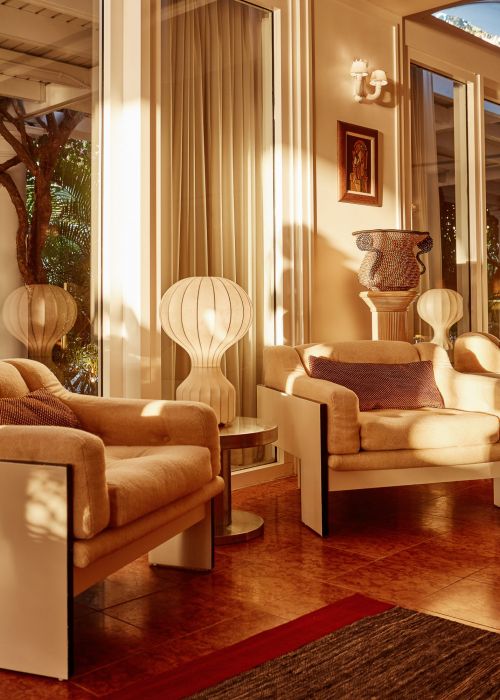 Living room interior with two beige armchairs, a small round side table, and decorative lamps, bathed in warm, inviting sunlight.