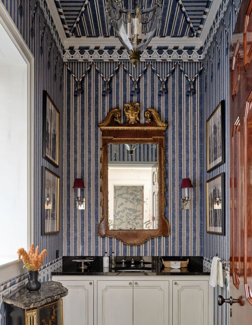 Ornate powder room interior with blue striped wallpaper, gilded mirror, white cabinets, and marble countertop, creating a luxurious design.