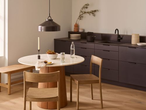 Interior of a modern dining room with a round marble table, wooden chairs and bench, and kitchen cabinets in the background.