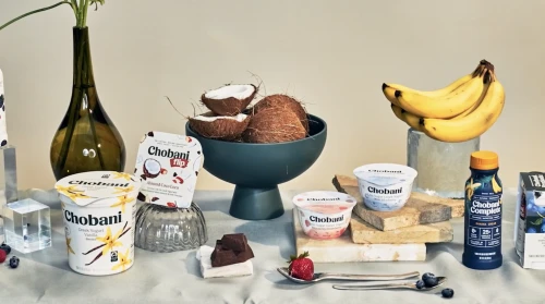 Still life with Chobani yogurt products, bananas, coconuts, and silver spoons, set on a white cloth. Food and beverage marketing photo.