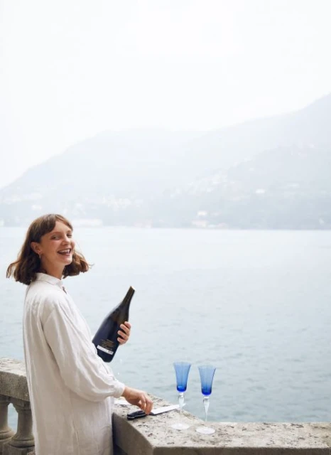 Woman in white dress smiles holding a champagne bottle outdoors near blue drinking glasses on a stone ledge with water and mountains.
