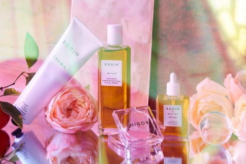 Still life of Rodin brand skincare products including creams, oils, and perfume arranged with peach roses and reflective surfaces.