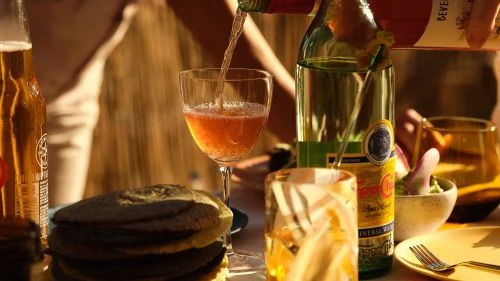 A glass is filled with a beverage next to a bottle on a table set with plates, food, and drinks, illuminated by warm lighting.