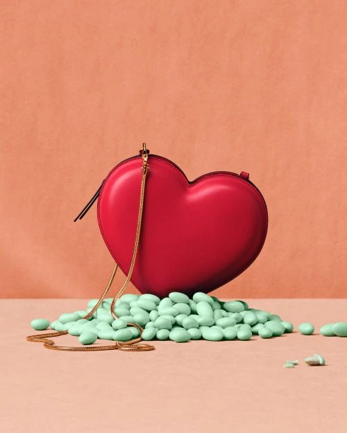 A heart-shaped red purse with a gold chain is surrounded by a pile of light green mints on a peach-colored surface in a studio setting.