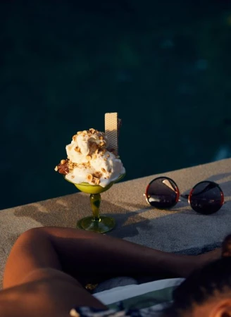 Ice cream sundae topped with nuts and a wafer alongside a pair of sunglasses on a ledge with a blurred background.