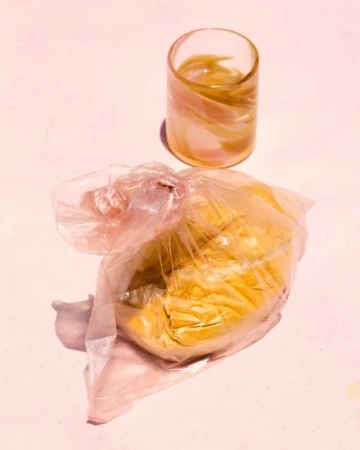 Still life photo of a glass of liquid and a yellow substance in a plastic bag sitting on a pink surface in a brightly lit indoor setting.