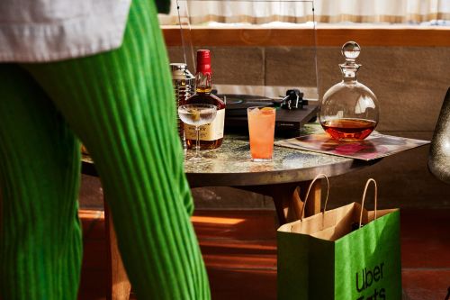 Cocktail drinks and record player on table with Uber Eats shopping bags, near a person wearing green pants in a warm, ambient indoor space.
