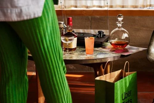 Cocktail drinks and record player on table with Uber Eats shopping bags, near a person wearing green pants in a warm, ambient indoor space.