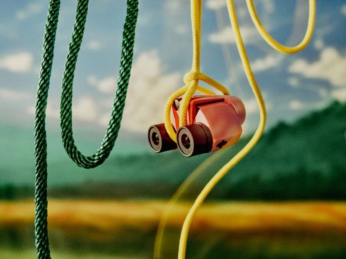 Close-up of toy binoculars hanging from yellow rope with green rope alongside, with a landscape background of fields and sky.