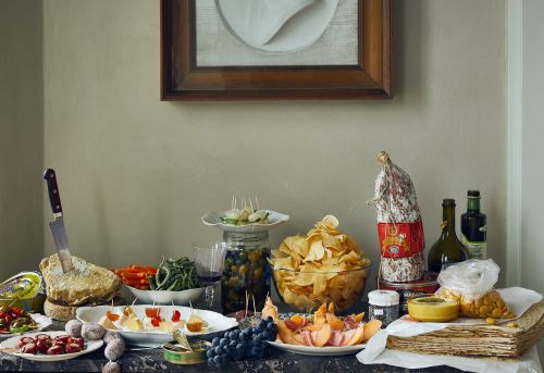 Still life of antipasto platter with salami, cheese, olives, chips, melon, crackers, and green beans on a dark marble counter.