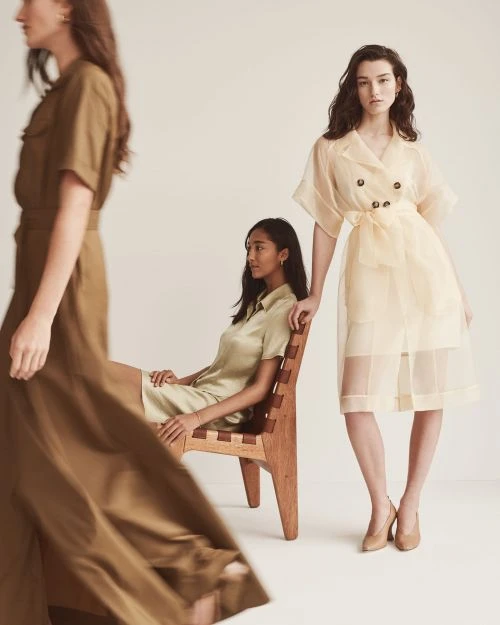 Three women in neutral-toned dresses pose in a studio. One woman sits in a wooden chair, while the other two stand. Fashion editorial.