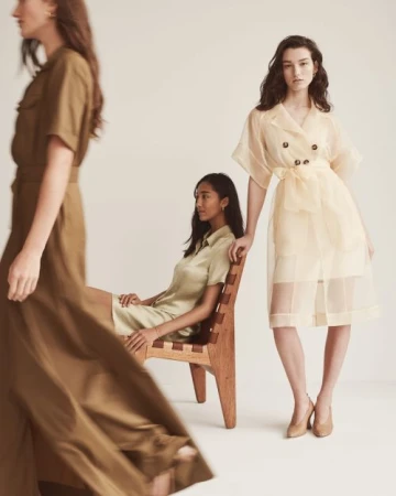 Three women in neutral-toned dresses pose in a studio. One woman sits in a wooden chair, while the other two stand. Fashion editorial.