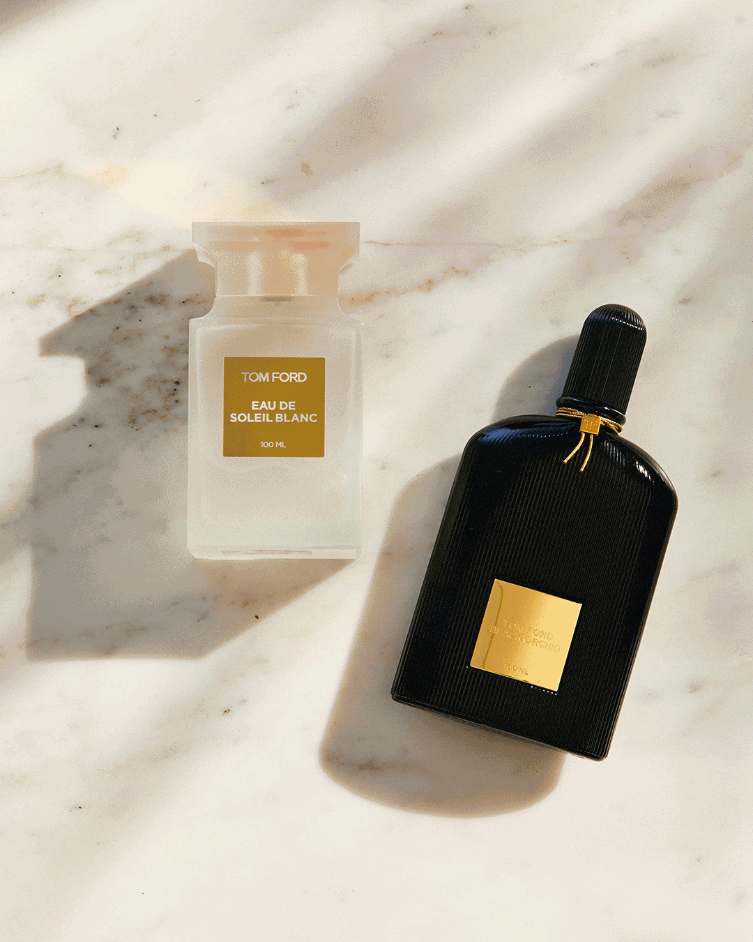 Two Tom Ford perfume bottles, one white and one black, sit on a textured marble surface with soft lighting and shadows.
