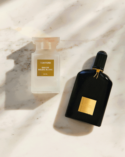 Two Tom Ford perfume bottles, one white and one black, sit on a textured marble surface with soft lighting and shadows.