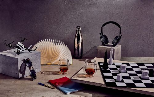 Still life composition featuring a drone, book lamp, chess set, glasses of whiskey, and headphones against a textured grey background.