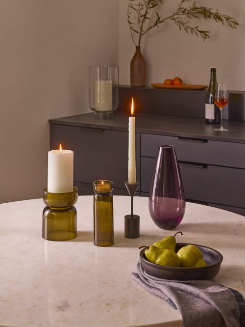Warm interior shot of candles burning on a marble table with green pears, a wine glass, a bottle of wine, and dark grey cabinets.