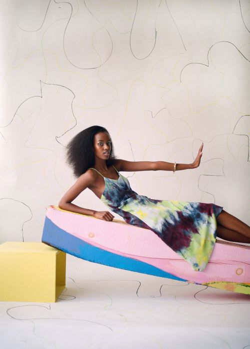 A young adult female poses in a vibrant tie-dye dress, reclining on colorful platforms in a studio setting, against a minimalist backdrop.