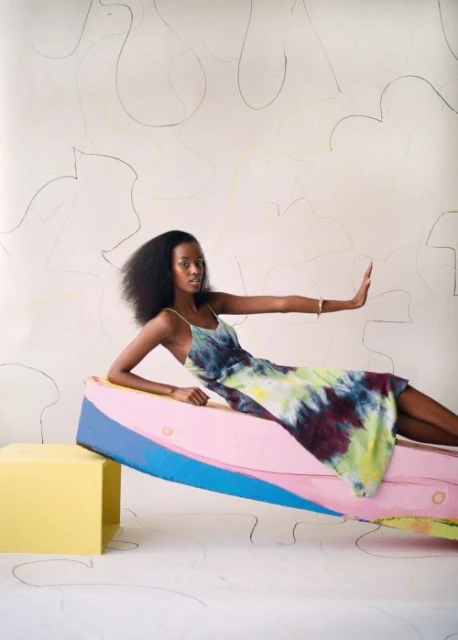 A young adult female poses in a vibrant tie-dye dress, reclining on colorful platforms in a studio setting, against a minimalist backdrop.