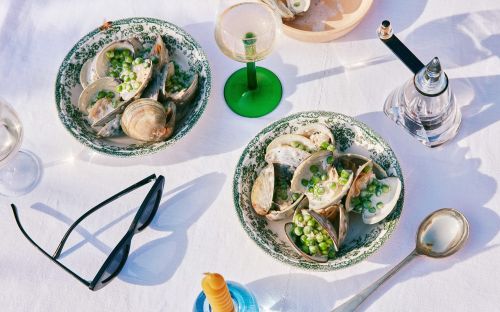 Overhead shot of two plates of clams with peas on a white tablecloth, accompanied by a pepper grinder, wine glass, spoon, candle, and sunglasses.