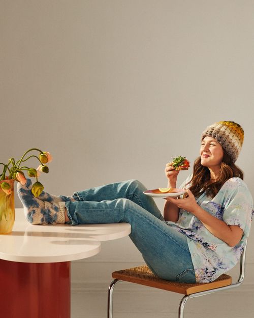 A smiling woman wearing a colorful beanie sits on a chair at a table, eating a sandwich with her feet up, in a naturally lit, indoor space.