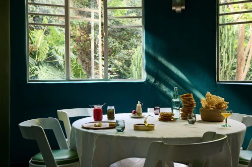 Indoor dining scene features a table set with dishes, food, and drinks, illuminated by natural light streaming through large windows.