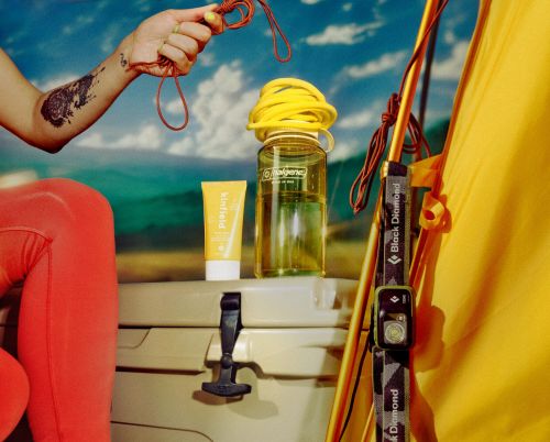 Outdoor gear and a water bottle are displayed on a cooler in front of a backdrop of a mountain lake under a bright blue sky.