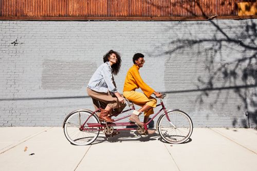 Two people, an adult female and an adult male, riding a tandem bicycle in an urban environment in the sunshine, with a brick wall background.