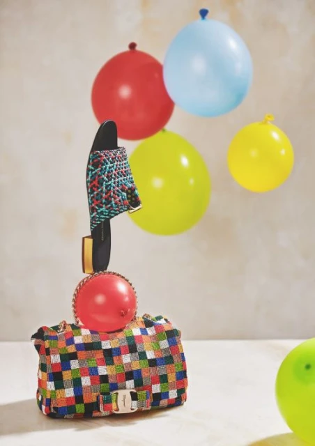 Still life composition featuring a woven purse, a slide sandal, and colorful balloons against a textured backdrop creating a playful scene.